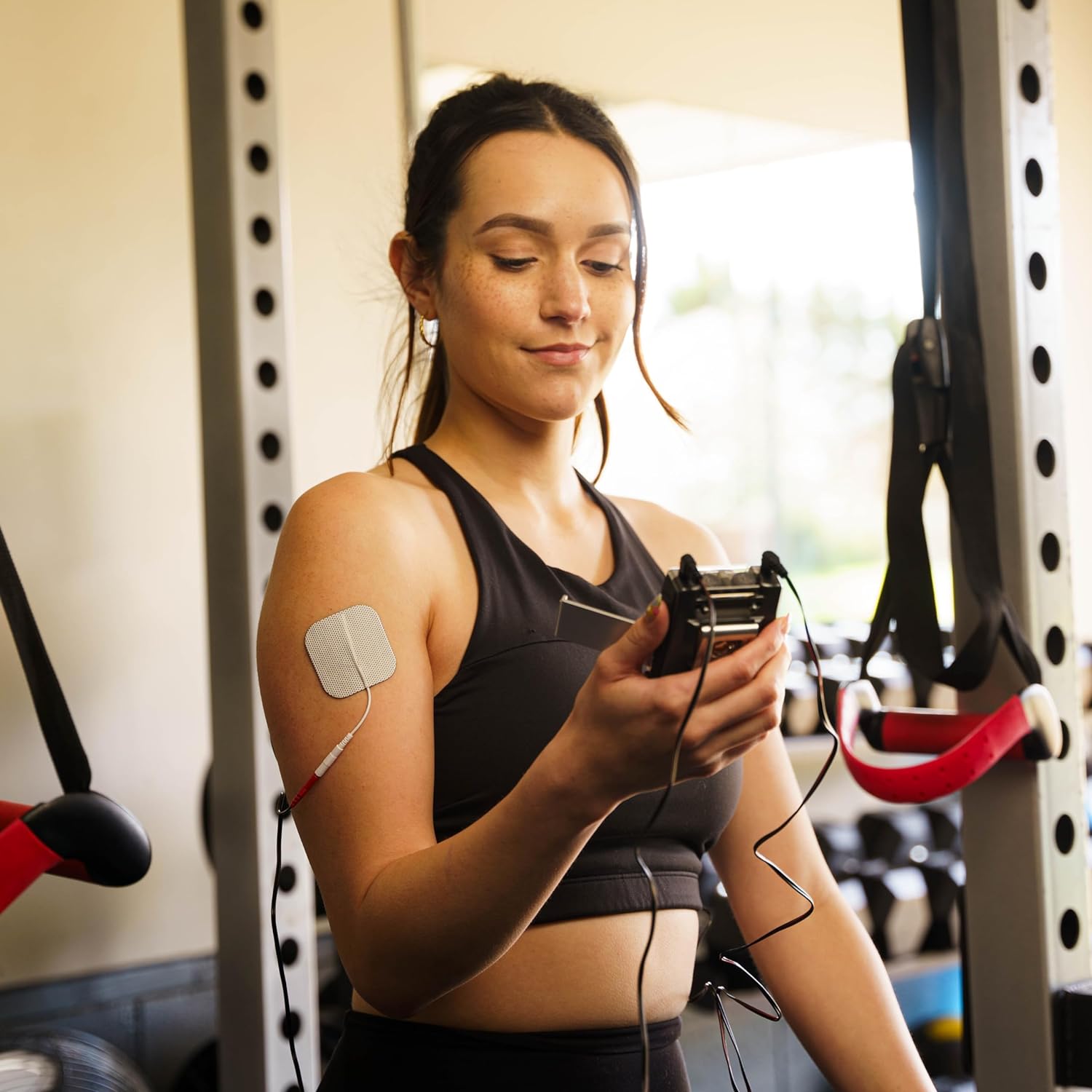 A woman holding a A gray TENS and EMS combo unit with electrodes on her shoulder