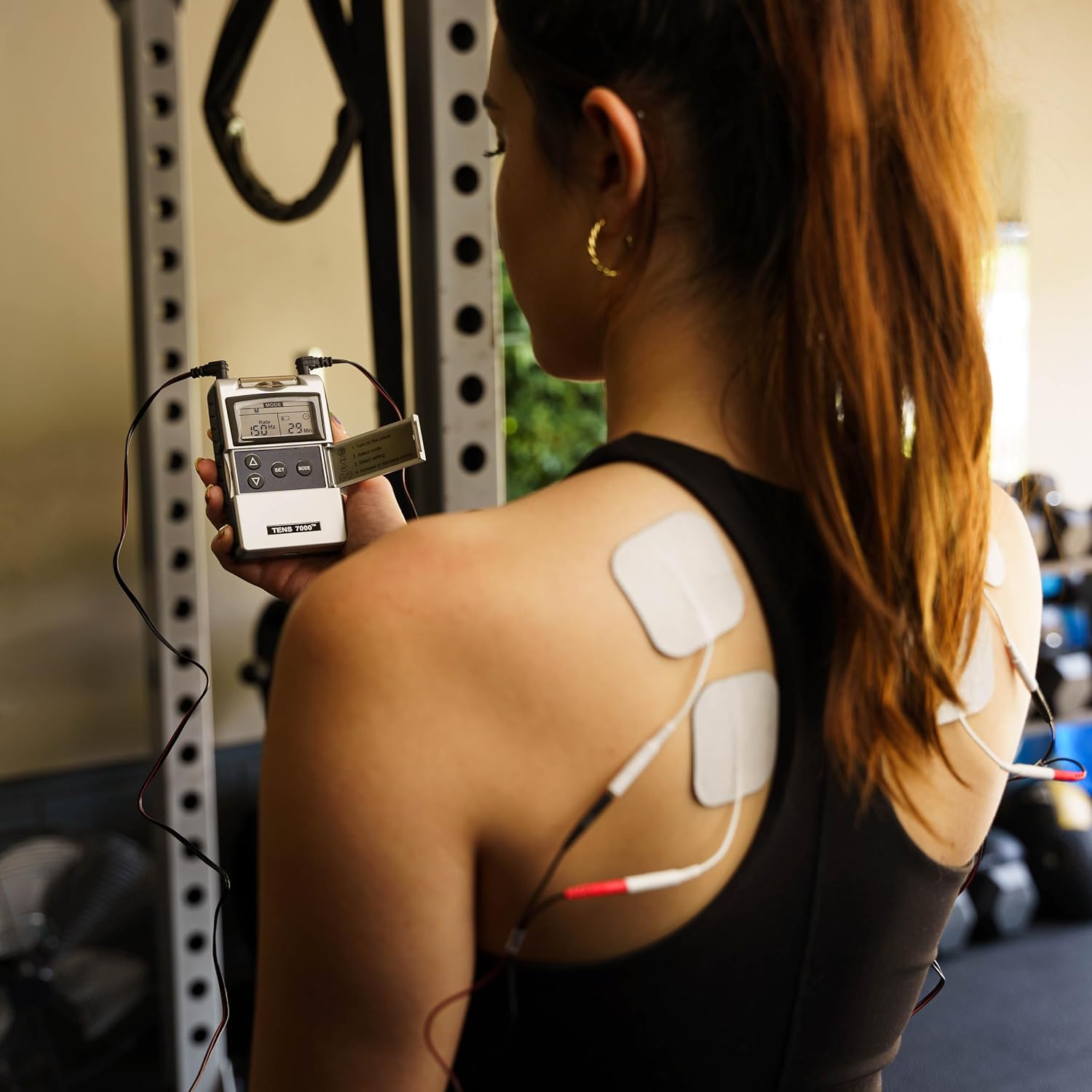 A woman in a gym holding a TENS unit with electrodes on her back