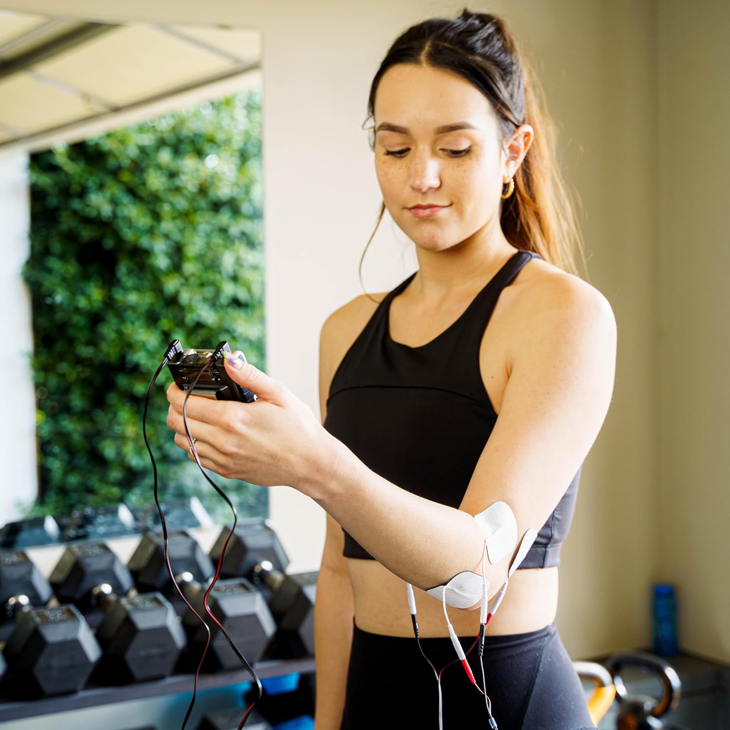 A woman holding a gray TENS and EMS combo unit with electrodes on her elbow