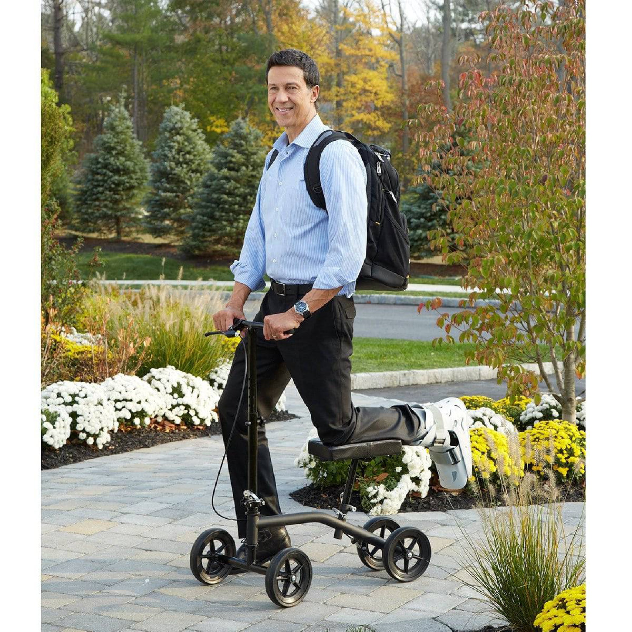 Man using a knee walker outdoors on a paved path with trees and flowers in the background