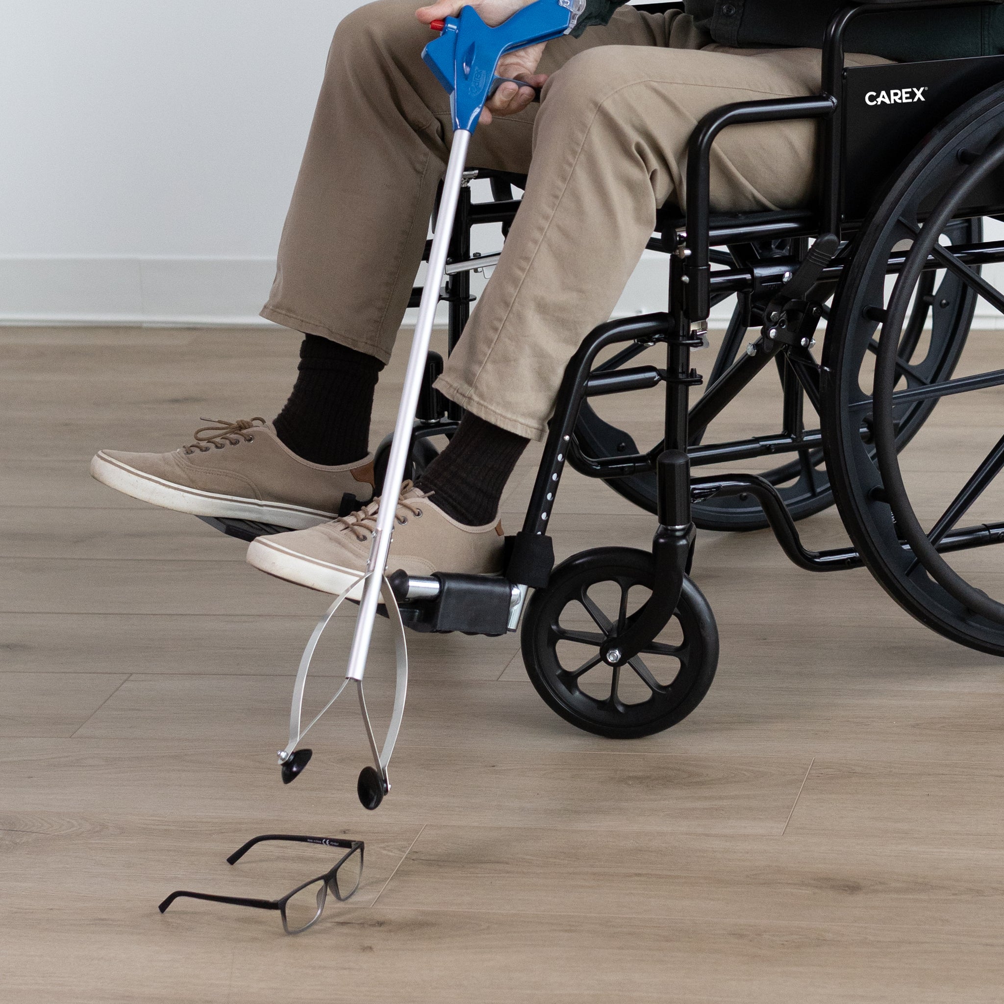 Person using a silver reacher grabber with a blue handle in a wheelchair, on a wooden floor.